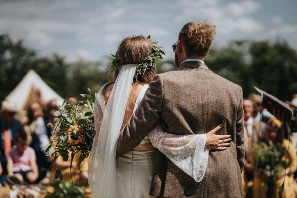 Couple standing together during an outdoor marquee wedding, with the bride in a floral crown and veil, holding a vibrant bouquet of sunflowers and greenery, and the groom wearing a tweed jacket, surrounded by guests and nature in the background.