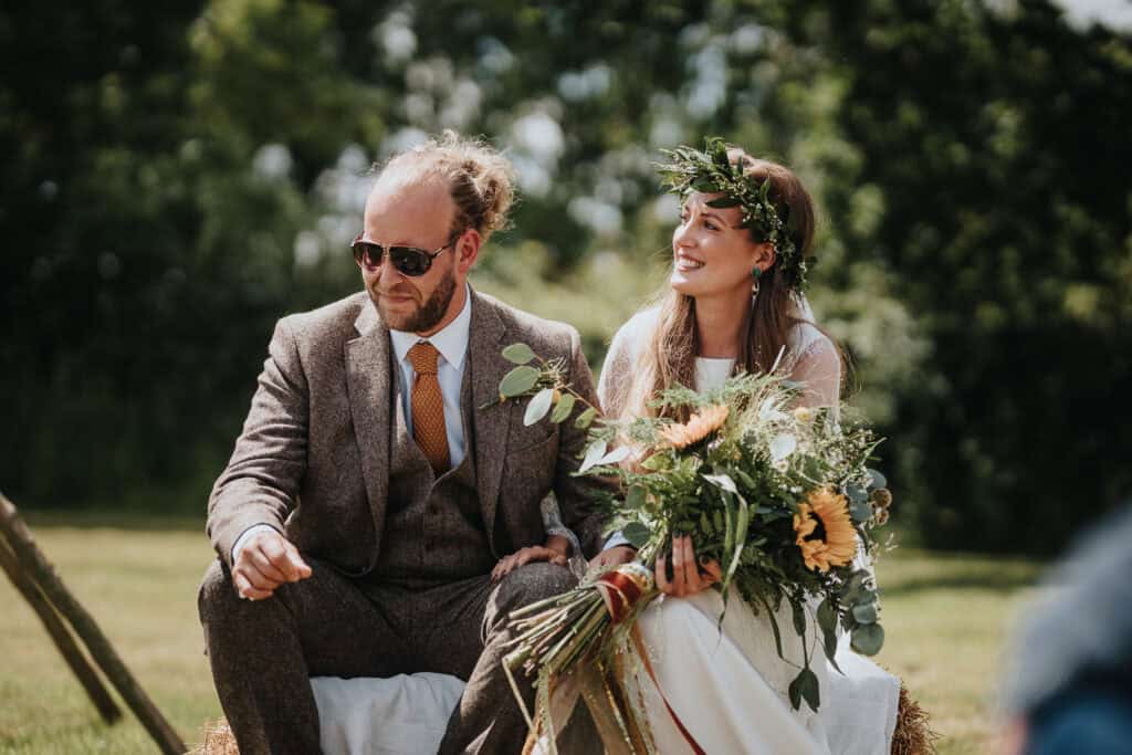 Outdoor summer farm marquee wedding featuring a bride with a floral crown and sunflower bouquet seated on a hay bale next to a groom in a tweed suit and sunglasses, enjoying the sunny countryside setting.