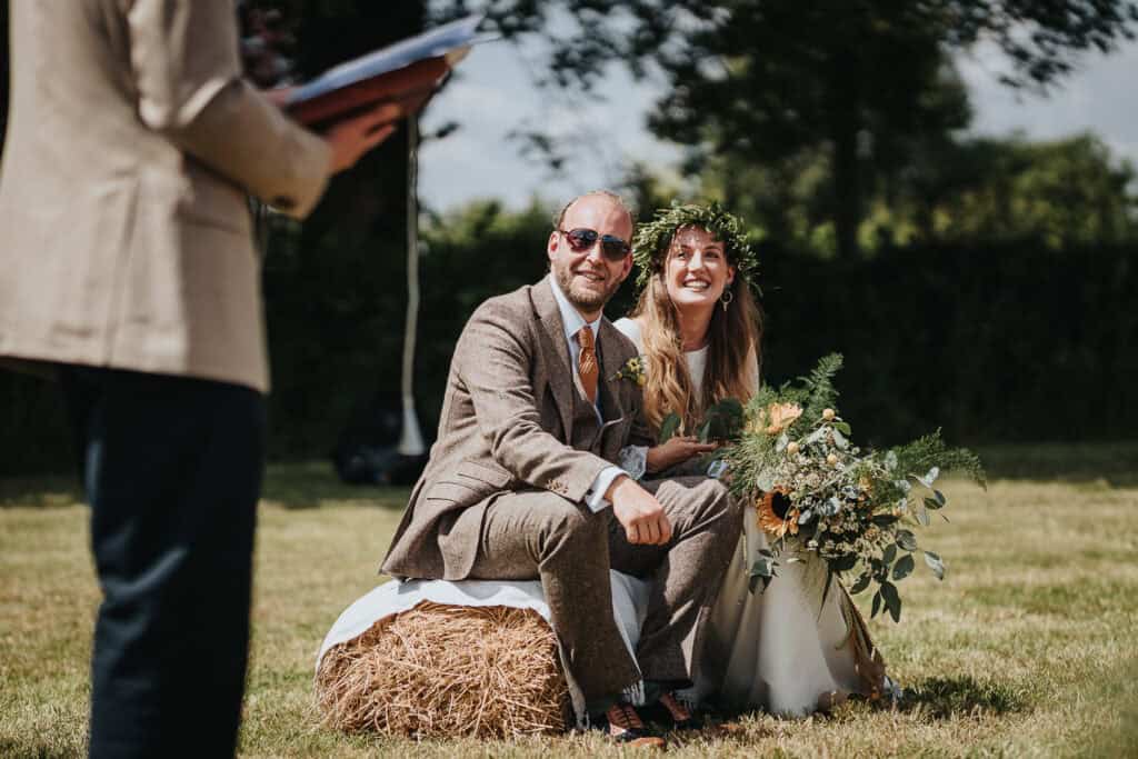 Outdoor rustic wedding ceremony with the bride and groom seated on a hay bale in a sunlit field. The groom wears a beige suit with sunglasses, while the bride smiles joyfully, holding a bouquet of greenery and sunflowers, adorned with a floral crown. A celebrant officiates in the foreground.