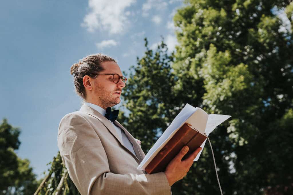 Wedding celebrant dressed in a light beige suit and bowtie, holding a book and conducting an outdoor ceremony. Set against a backdrop of lush green trees and a bright blue sky, the scene exudes elegance and charm, capturing the essence of a beautiful outdoor wedding.