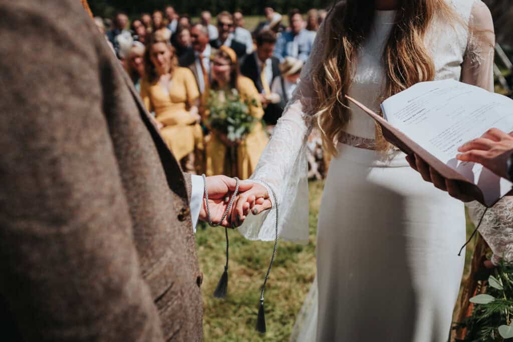 An intimate outdoor wedding ceremony in a lush green setting, featuring a couple exchanging vows and holding hands, with close friends and family gathered in the background. The bride is wearing a beautiful long-sleeve gown, while the officiant reads from a book.