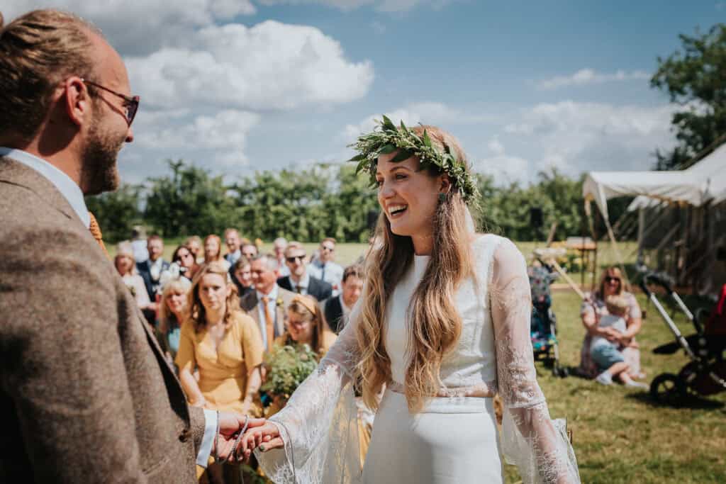 A joyful outdoor marquee wedding ceremony in a sunny field, featuring a bride with a floral crown and lace dress smiling at her groom in a tweed suit. Guests are seated on benches in the background under a bright blue summer sky with fluffy white clouds.