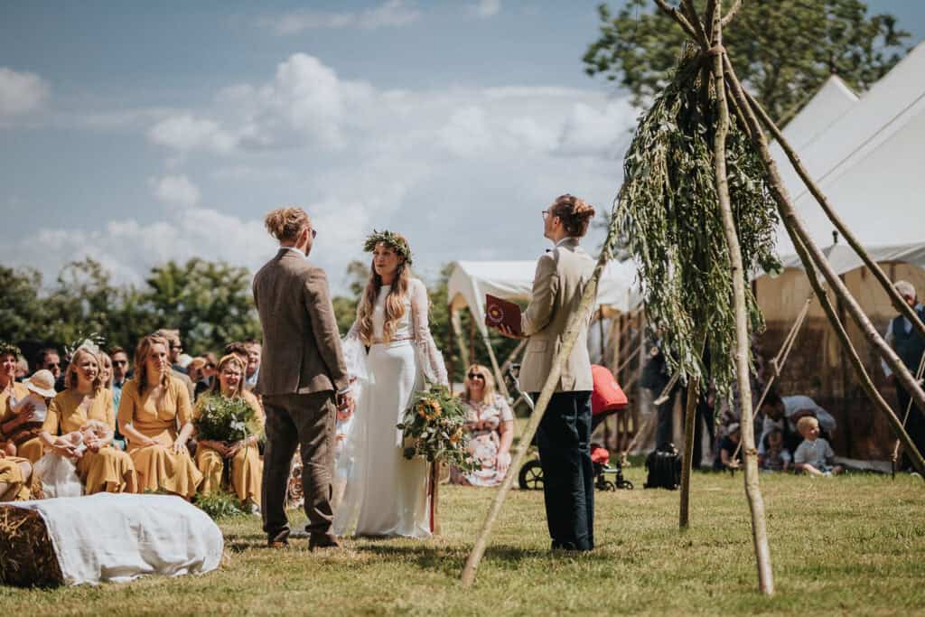 Outdoor wedding ceremony on a sunny day featuring a couple standing under a rustic floral arch with a celebrant officiating. Guests are seated on hay bales and dressed in cheerful tones, with a marquee tent visible in the background, creating a charming countryside setting.