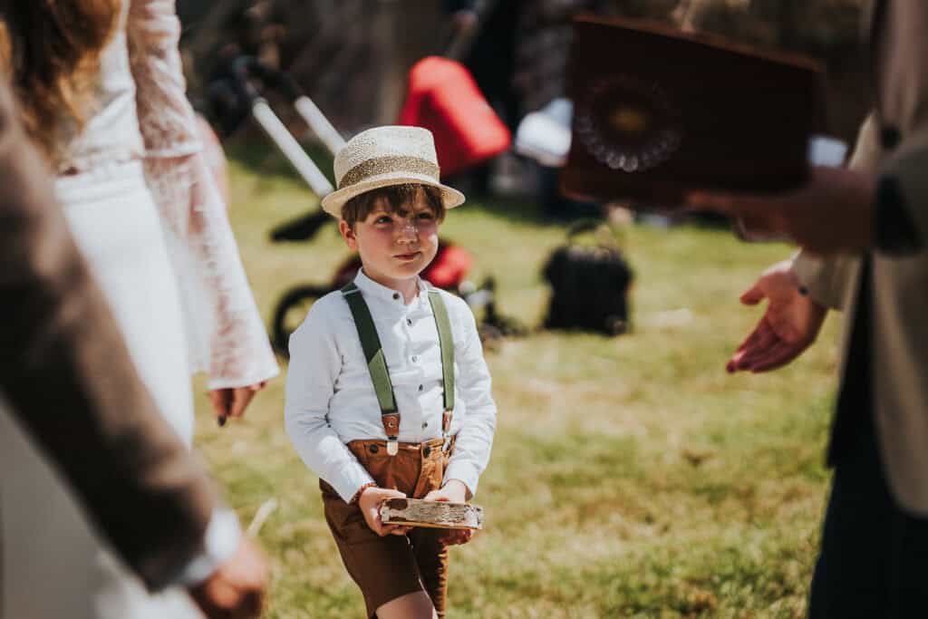 Adorable young ring bearer dressed in a white shirt, brown shorts with green suspenders, and a straw hat, holding the wedding rings in a rustic outdoor setting.