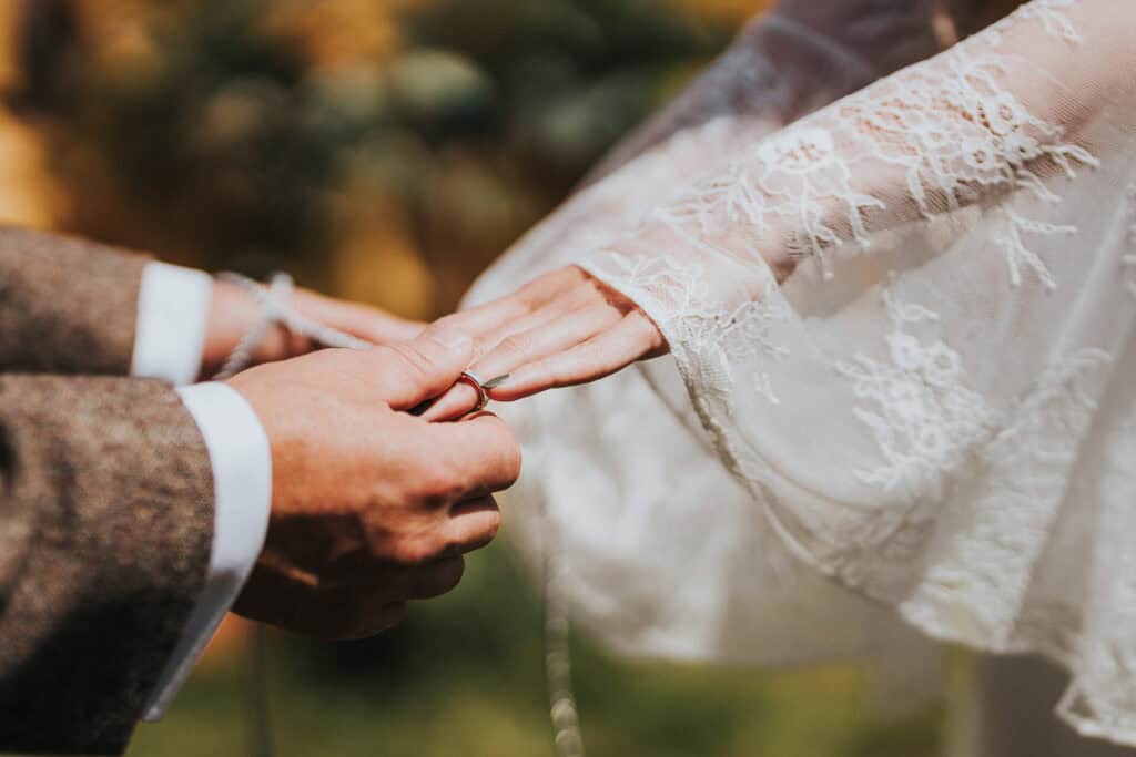 Close-up of a bride and groom exchanging wedding rings during an outdoor ceremony, with the bride's lace sleeve and the groom's tweed suit visible.