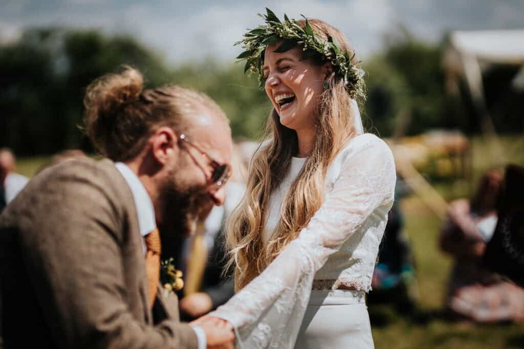 During their outdoor wedding, the groom struggles to put the ring on the bride's finger, leading to a lighthearted and laughter-filled moment between the couple, captured in the midst of a beautiful, sunny ceremony.