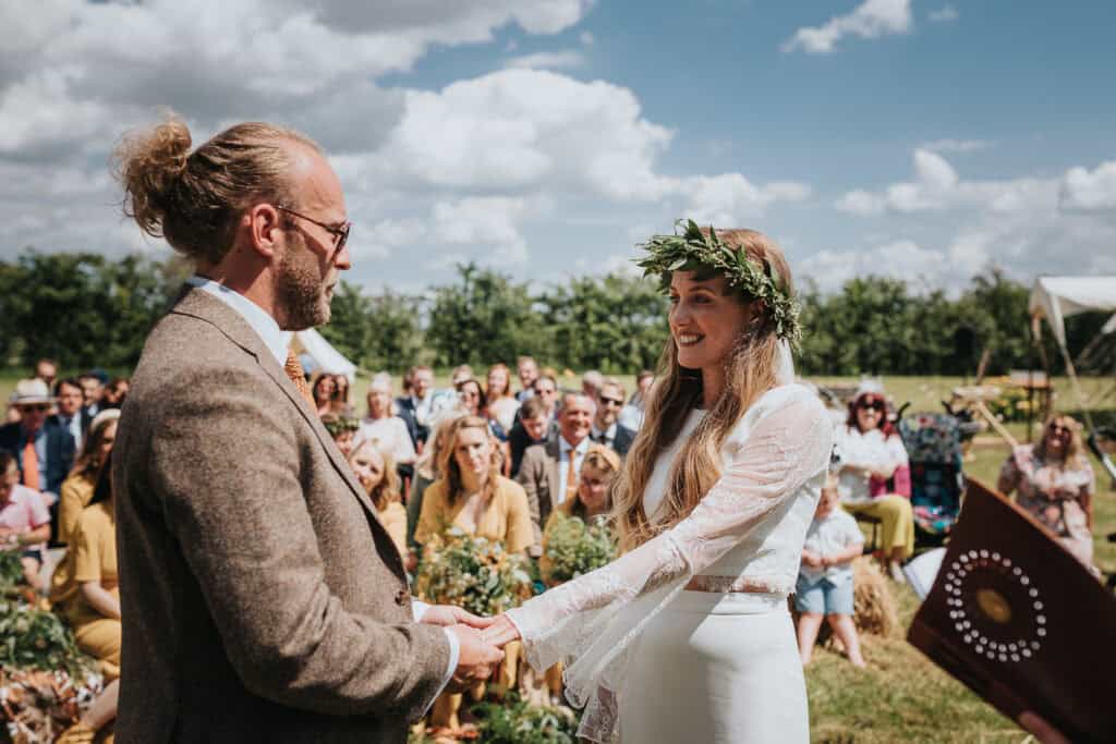 Outdoor wedding ceremony in a scenic countryside setting with a couple exchanging vows. The bride, wearing a long-sleeved white gown and a flower crown, smiles warmly at the groom, dressed in a brown tweed suit. The event is surrounded by greenery, with family and friends seated on hay bales, enjoying the sunny day under a bright blue sky with fluffy clouds.