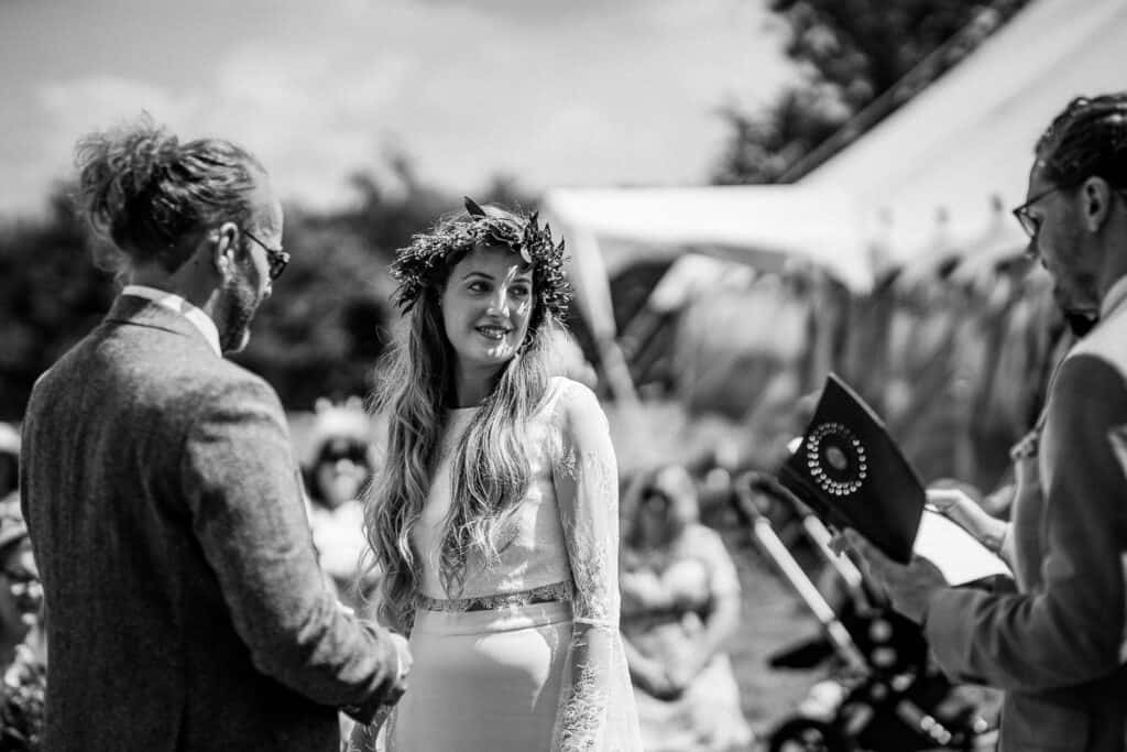 Black-and-white photograph of an outdoor wedding ceremony featuring a bride wearing a floral crown and lace gown, standing beside the groom in a tailored suit. A celebrant holds an open booklet while officiating the ceremony, with a marquee tent and guests visible in the background.