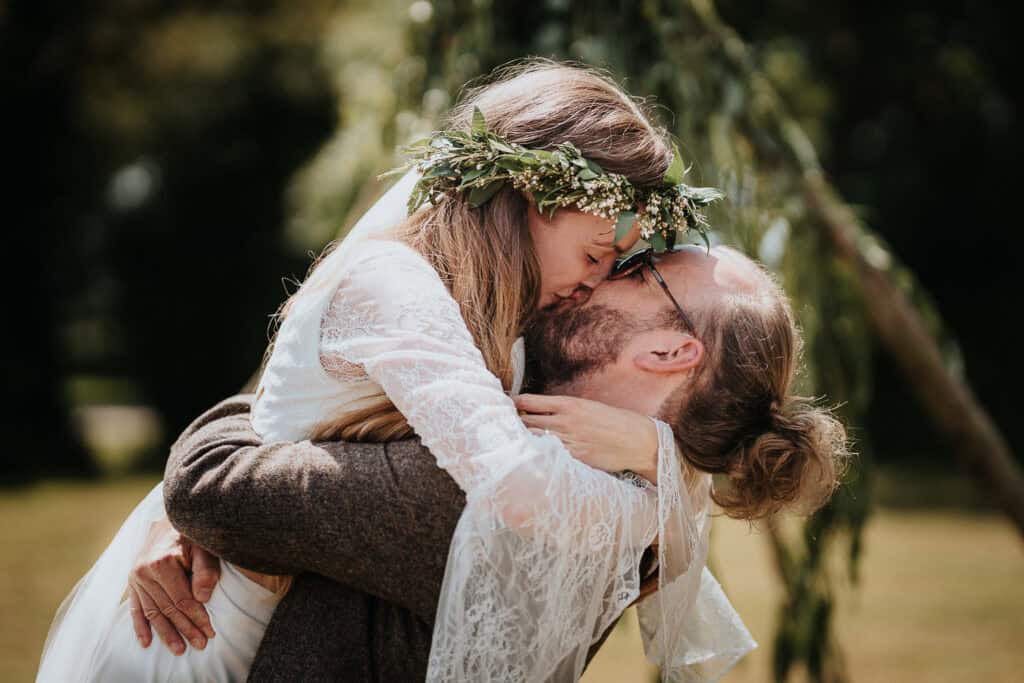 First kiss captured in a rustic outdoor wedding, with the bride wearing a lace gown and floral crown, embraced by her partner beneath a serene backdrop of greenery.
