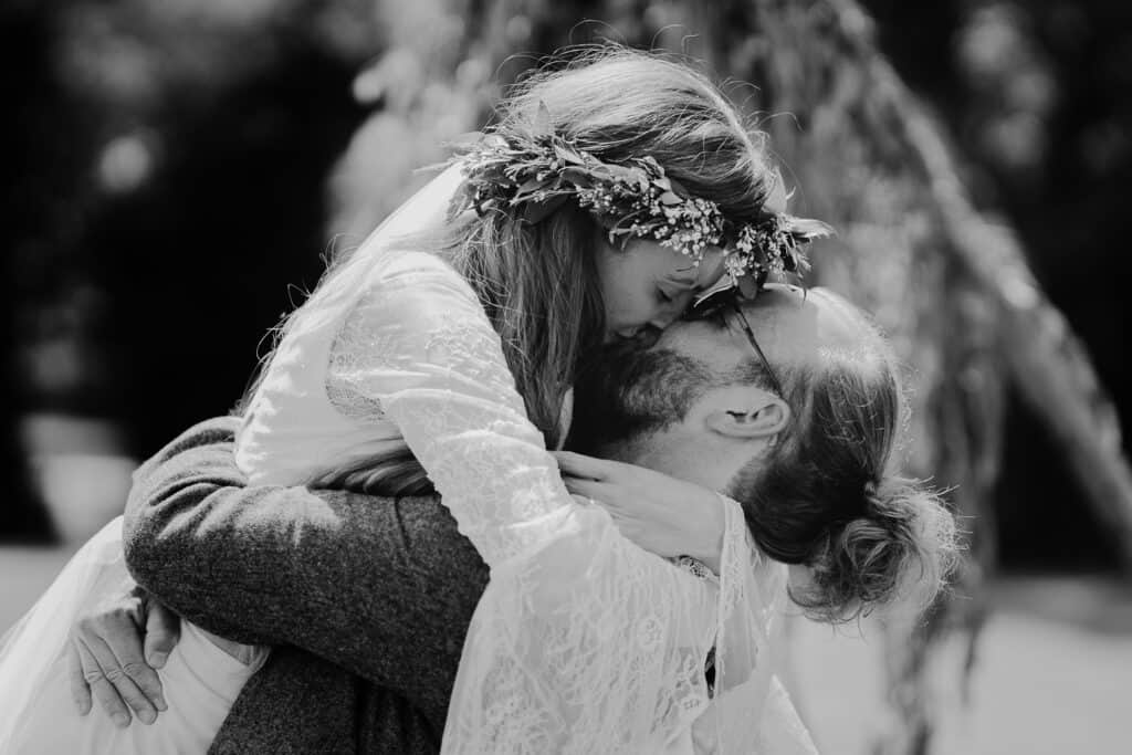 Black and white photo capturing a tender first kiss between a newlywed couple after their farm wedding ceremony. The bride wears a lace dress and a floral crown, while the groom lovingly embraces her outdoors in a rustic, natural setting.