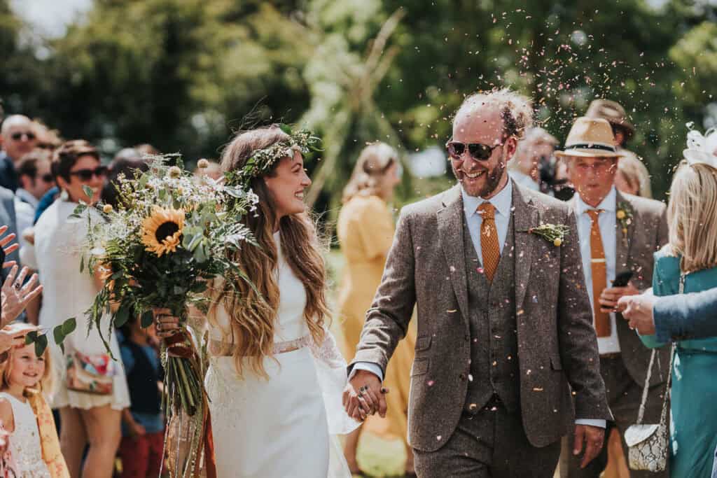 Joyful couple walking hand in hand at their outdoor marquee wedding, surrounded by guests showering them with confetti. The bride holds a vibrant bouquet with sunflowers and greenery, while the groom beams in a classic tweed suit with an orange tie.