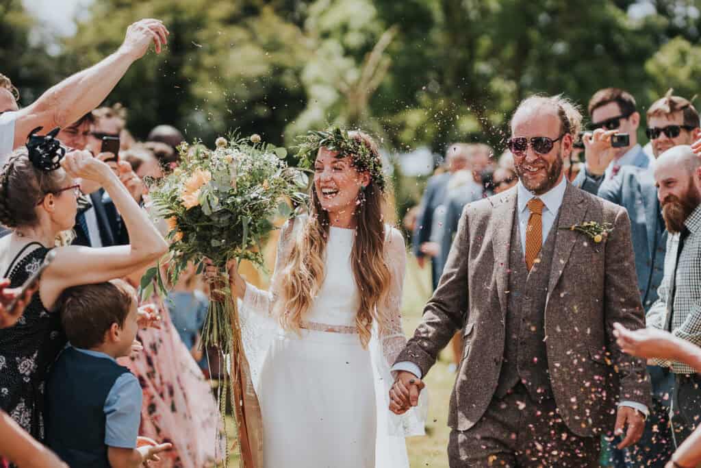Wedding couple walking hand in hand outdoors under a shower of confetti, surrounded by joyful guests celebrating. The bride, wearing a white gown with floral headpiece, smiles radiantly while holding a lush bouquet. The groom, in a stylish brown tweed suit, looks confident and happy. A vibrant and heartwarming moment captured in natural light amidst a green countryside setting.