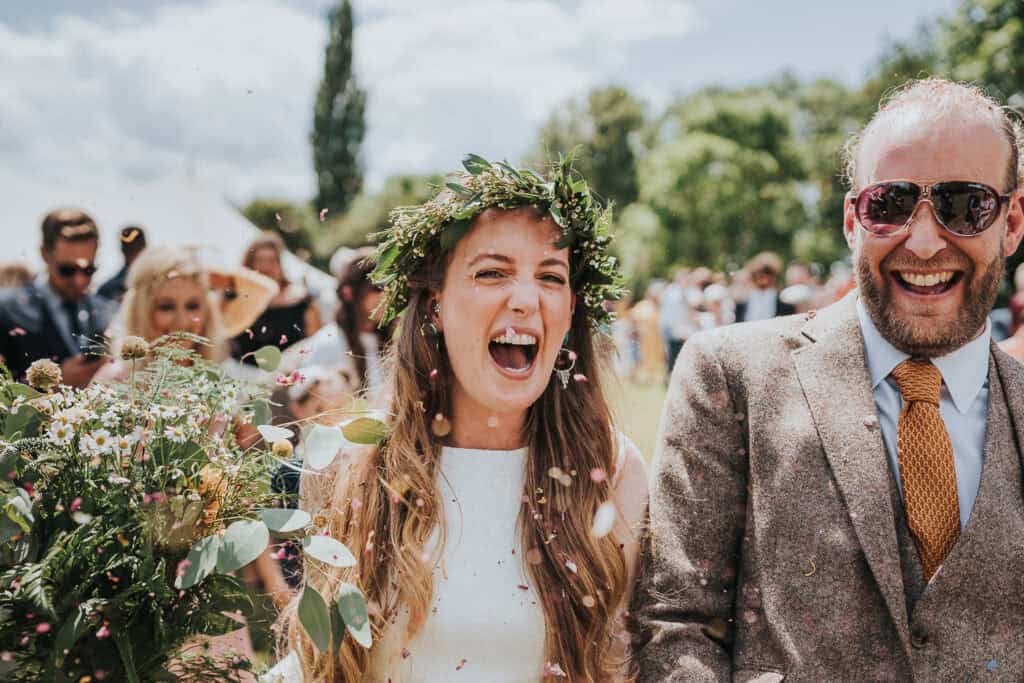 Joyful wedding moment captured outdoors under a clear sky, featuring a radiant bride wearing a flower crown and holding a bouquet, laughing alongside a smiling man in a brown tweed suit and orange tie. Guests in the background celebrate with confetti, adding vibrancy to the rustic garden wedding scene.