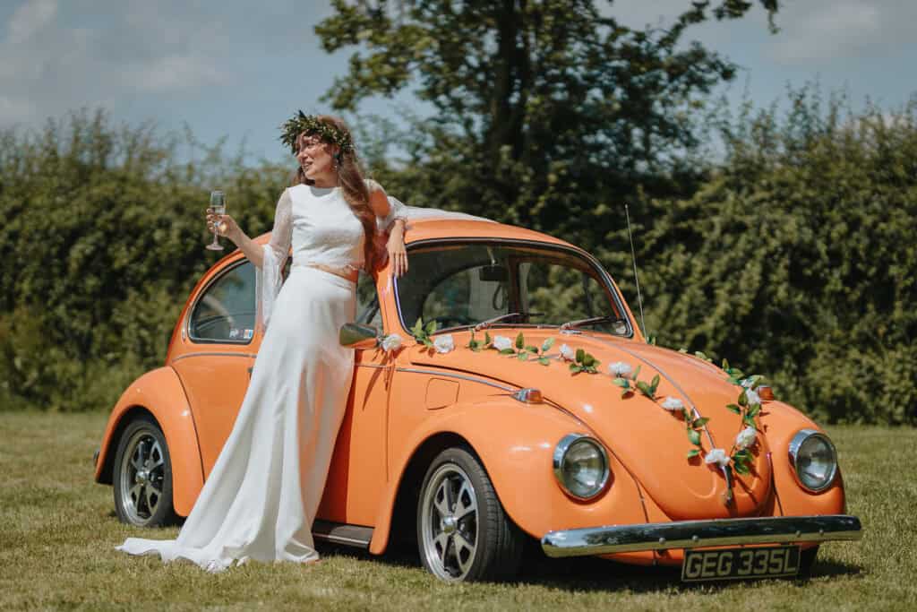 Rustic wedding photography featuring a bride in a flowing white gown and floral crown, leaning against a vintage orange Volkswagen Beetle adorned with floral garlands, set in a scenic countryside field with lush greenery in the background.