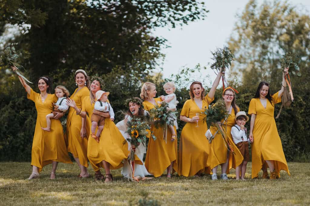 Group of bridesmaids wearing matching yellow dresses, holding floral bouquets, and posing outdoors with children at a farm-style wedding. The scene is set in a lush green garden under natural light, creating a joyful and rustic vibe.
