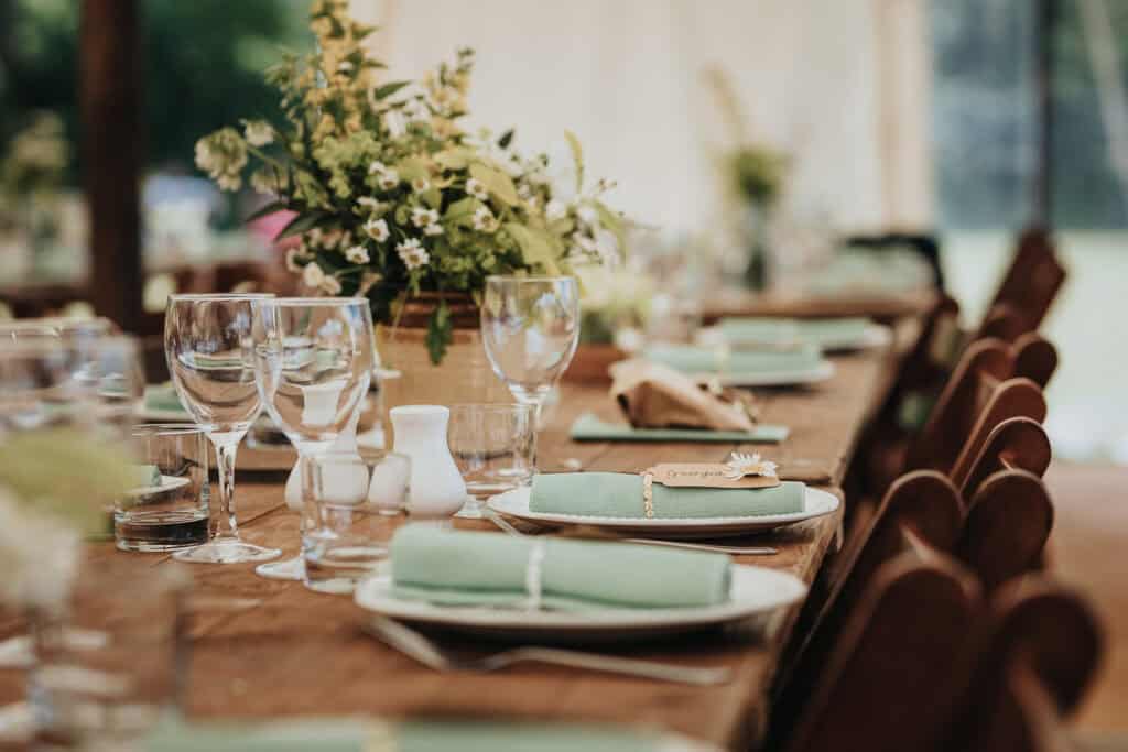 Rustic wedding table setting featuring wooden tables adorned with green napkins, glassware, and floral centrepieces under a marquee tent.