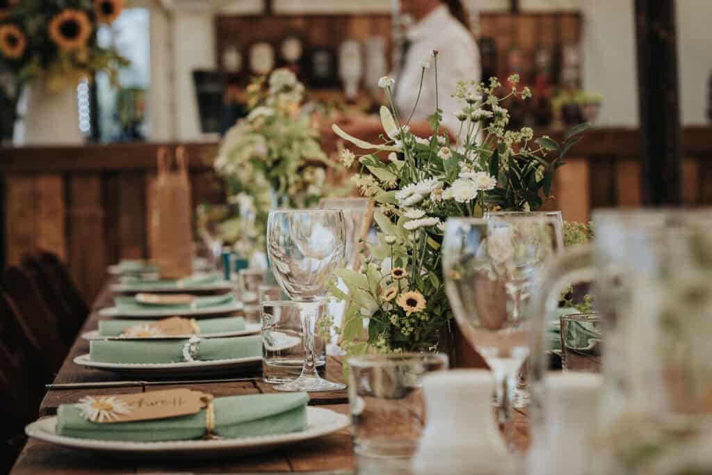 Rustic wedding table setting featuring green napkins, floral centrepieces with wildflowers, and wooden accents in a cozy farm-style venue.