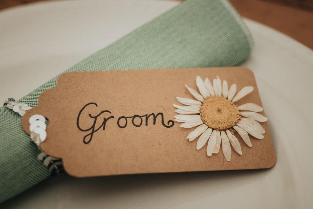 Rustic wedding table place card for the groom, featuring a handcrafted brown tag with elegant calligraphy and a dried daisy flower, placed atop a folded green napkin on a white plate.