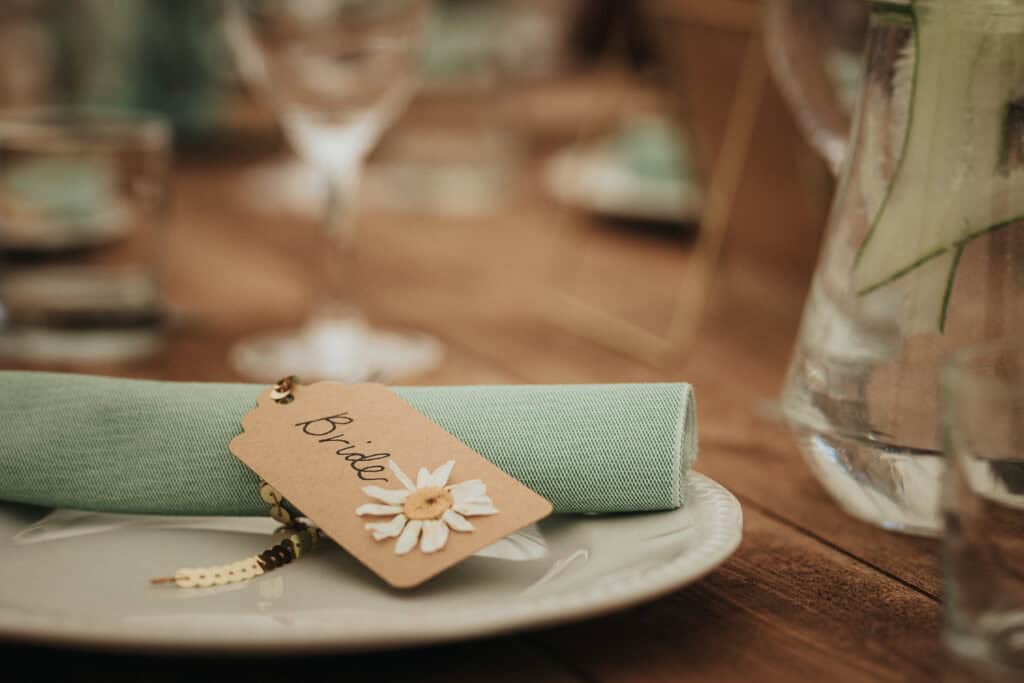 Elegant place setting for a wedding, featuring a light green napkin neatly folded on a white plate with a handcrafted tag labeled 'Bride,' adorned with a small flower design, set on a rustic wooden table.