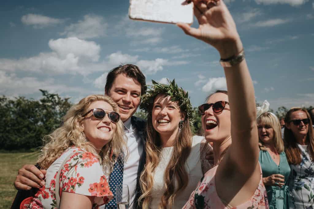 A cheerful outdoor wedding selfie featuring the bride in a white dress and a greenery crown, surrounded by her bridesmaids and brother. Everyone is smiling and enjoying the moment, with guests and a sunny green landscape in the background.