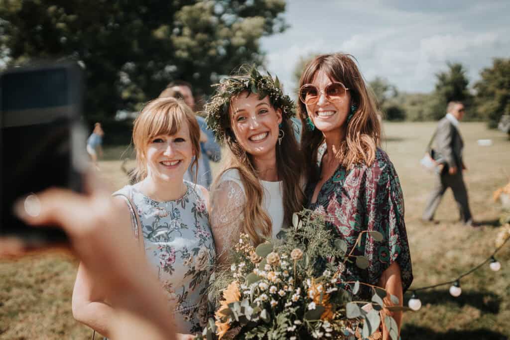 A joyful outdoor wedding moment featuring the bride in the centre, wearing a floral crown and holding a bouquet of wildflowers, smiling warmly at the camera. She is accompanied by two women on either side, dressed in floral and patterned outfits, sharing her happiness. The scene is set in a sunny field with a rustic and relaxed wedding vibe, with trees and blurred guests in the background.