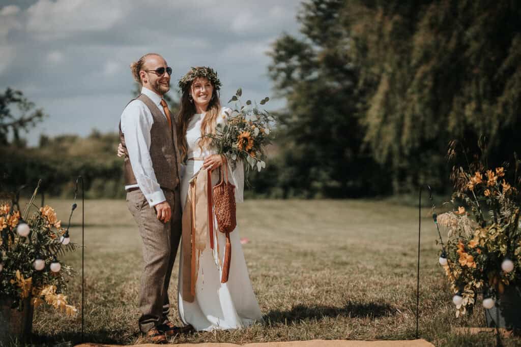 A couple standing outdoors on a grassy field during a rustic wedding ceremony, surrounded by floral arrangements. The bride is wearing a flowing white dress with a floral crown, holding a bouquet, and the groom is dressed in a beige vest and trousers with a white shirt and tie. The setting features a natural and bohemian vibe with soft greenery and warm-toned flowers under a sunny sky.