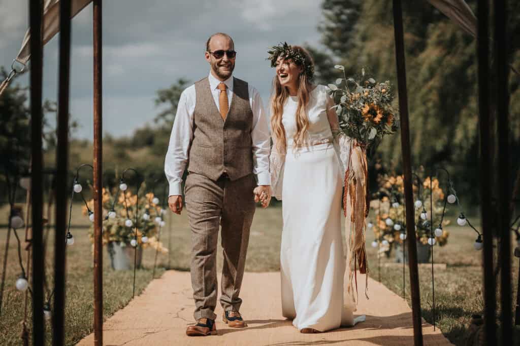 Rustic outdoor wedding breakfast entrance with the bride and groom walking hand in hand. The bride is in a flowing white dress with a floral crown and holding a bouquet of sunflowers and greenery, while the groom wears a beige vest and tie. The pathway is lined with floral arrangements, leading to a beautifully decorated garden setting for the wedding breakfast.