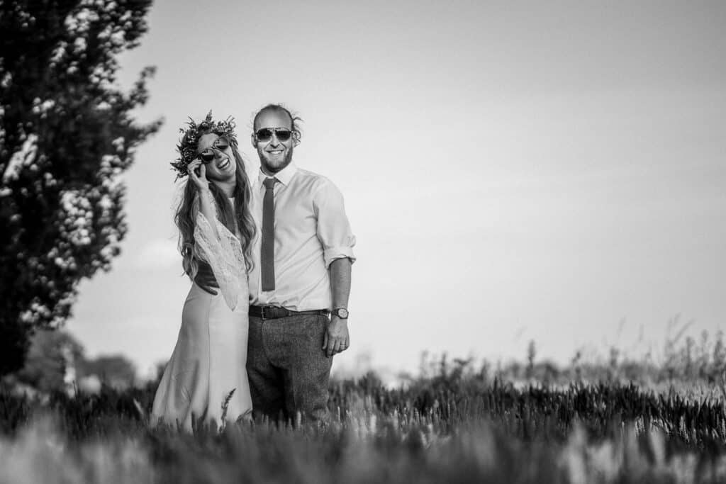 A black-and-white wedding portrait of a bride and groom standing in a field. The bride is wearing a long wedding dress and a floral crown, smiling joyfully, while the groom is dressed in a shirt and tie with sunglasses, also smiling. The background features trees and an open countryside landscape.