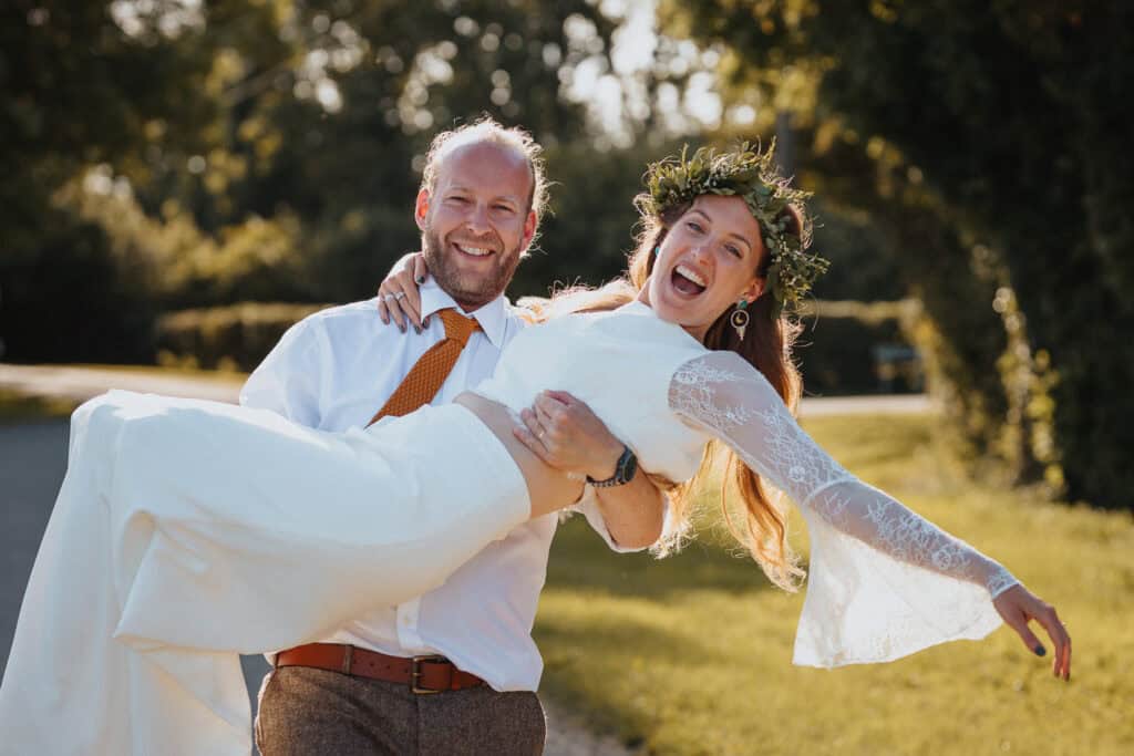 Joyful outdoor wedding photo of a bride and groom during their portrait session, with the groom playfully holding the bride in his arms. The bride, wearing a beautiful two-piece dress and a floral crown, beams with laughter, while the groom smiles warmly in a white shirt and orange tie. Captured in a sunny, natural setting with lush greenery in the background.