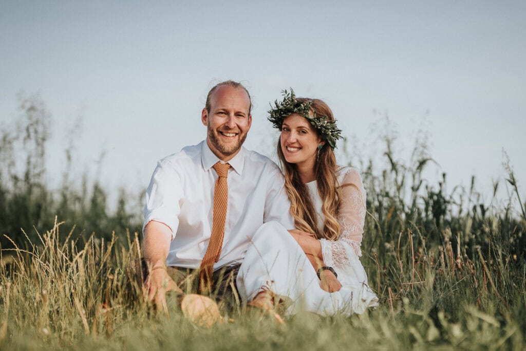 Outdoor wedding portrait of a bride and groom sitting together in a serene meadow, with the bride wearing a floral crown and a lace dress, and the groom dressed in a white shirt and tie, both smiling warmly under a clear blue sky.