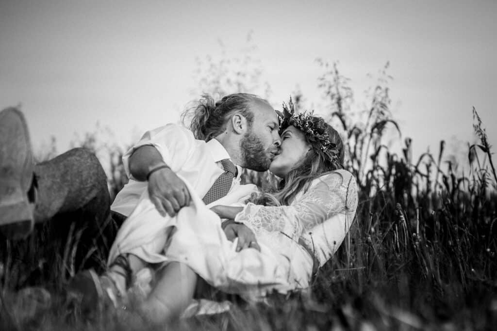 Black and white photo of a bride and groom sharing a romantic kiss while sitting in a grassy field. The bride wears a floral crown and lace wedding dress, while the groom is dressed in a white shirt and tie. The serene natural backdrop adds a timeless and intimate feel.