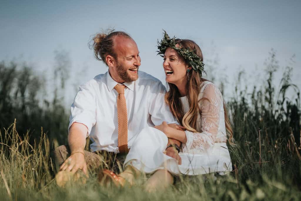 An intimate moment captured between a bride and groom sitting together in a lush meadow, laughing joyfully, with natural greenery and a serene sky in the background. The bride wears a floral crown and a white dress, while the groom is dressed in a white shirt and tie, radiating happiness and love.