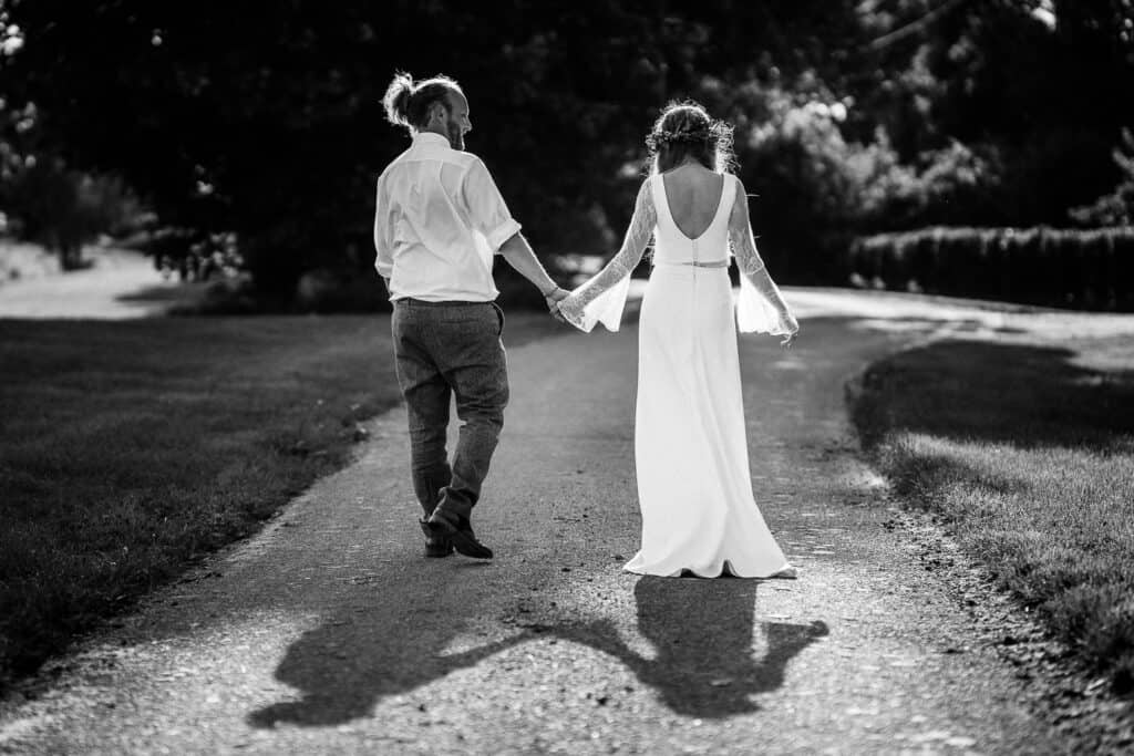 Black and white photograph of a bride and groom walking hand in hand along a sunlit path, their elongated shadows stretching out behind them. The bride wears a flowing white gown, and the groom is dressed in a casual shirt and trousers, with trees and greenery in the background.
