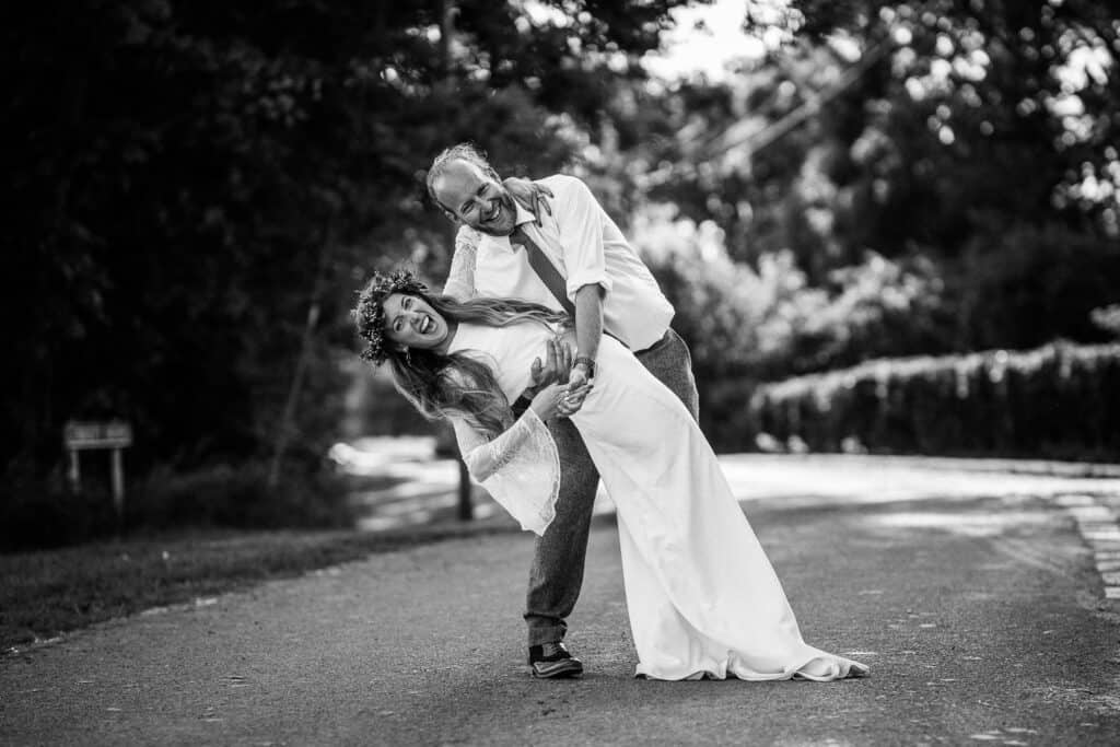 Black and white photograph of a bride and groom posing playfully on a quiet country road, with the groom leaning backward and the bride laughing as she leans forward in her elegant wedding dress and flower crown. Surrounded by trees and natural scenery, the image captures a joyful and candid moment of their special day.