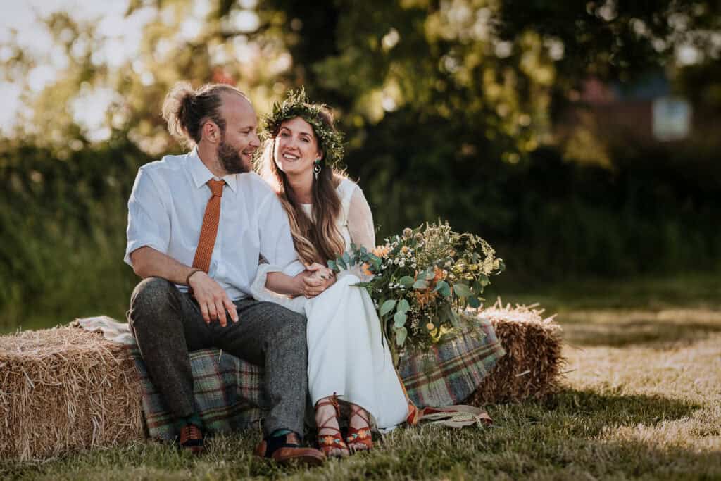 Bride and groom sitting on a hay bale during golden hour, surrounded by a rustic outdoor setting, with the bride holding a vibrant bouquet and wearing a floral crown, and the groom in a white shirt and orange tie, sharing a joyful moment together.
