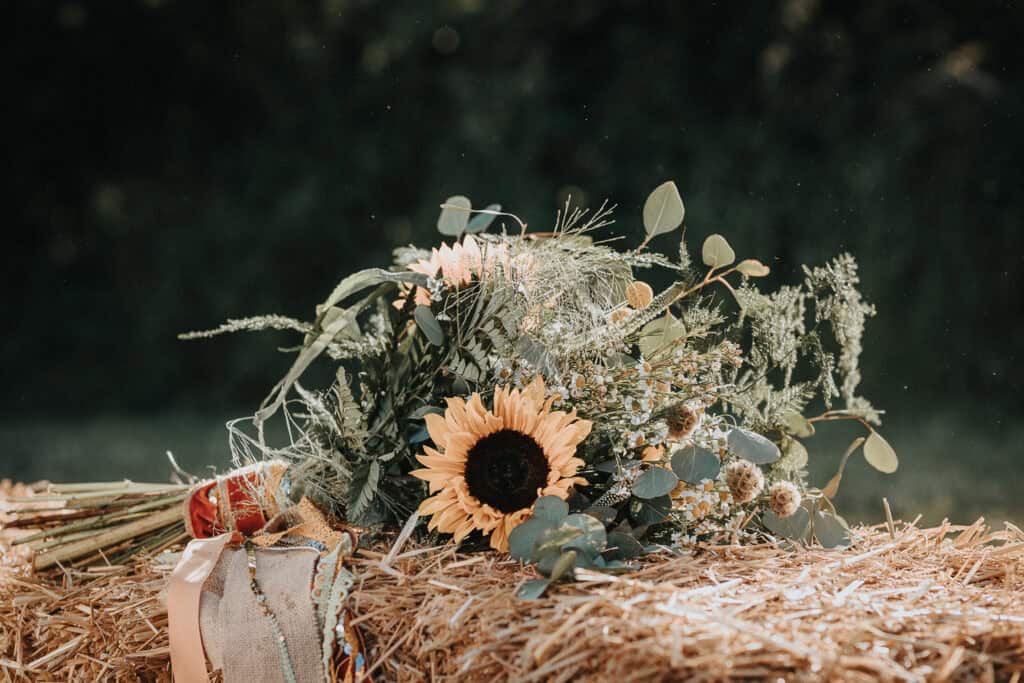 Rustic bouquet featuring a vibrant sunflower, greenery, and wildflowers, resting on a straw bale, with a natural outdoor backdrop.