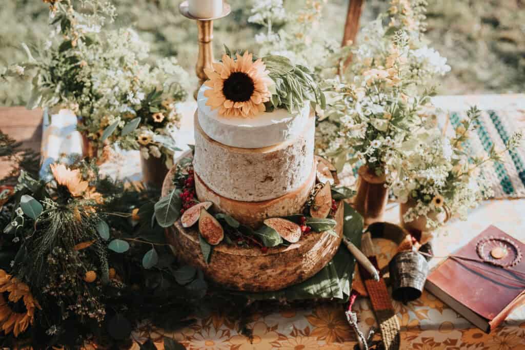 Rustic sunflower-themed wedding cheese cake table adorned with fresh greenery, figs, and floral arrangements, featuring a two-tier cheese cake topped with a sunflower and surrounded by a natural woodland-inspired setting.