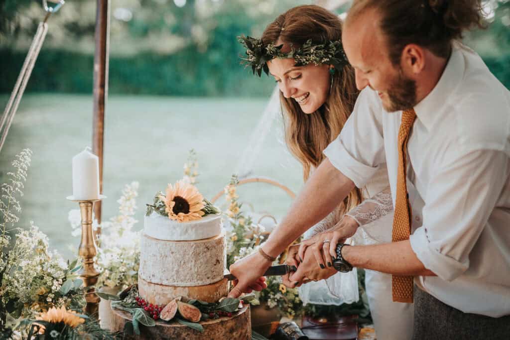 Rustic outdoor wedding featuring a smiling bride and groom cutting a beautiful tiered cake adorned with sunflowers and greenery, set amidst a picturesque natural backdrop.