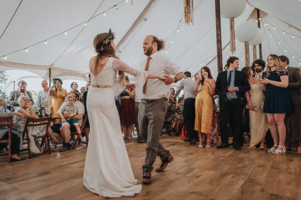 Bride and groom sharing their first dance under a charming marquee, surrounded by guests and illuminated by soft string lights, creating a romantic and rustic wedding atmosphere.