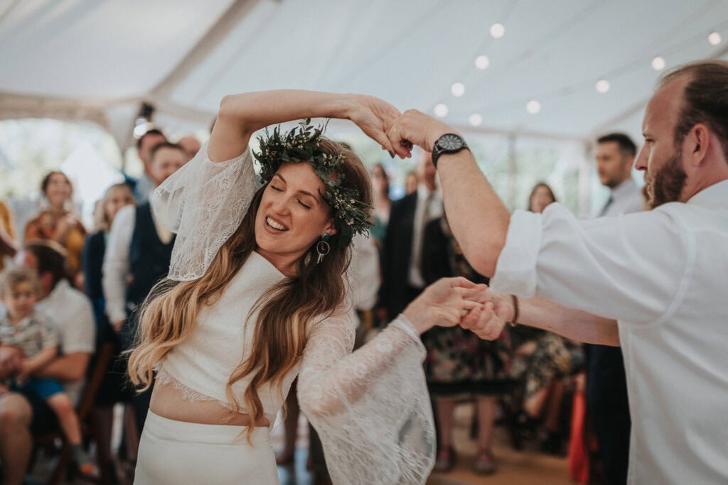 An enchanting moment of a bride and groom sharing their first dance under a beautifully lit marquee, surrounded by joyous family and friends. The bride, wearing a delicate two-piece lace gown and a floral crown, gracefully twirls with her partner, capturing the essence of love and celebration.