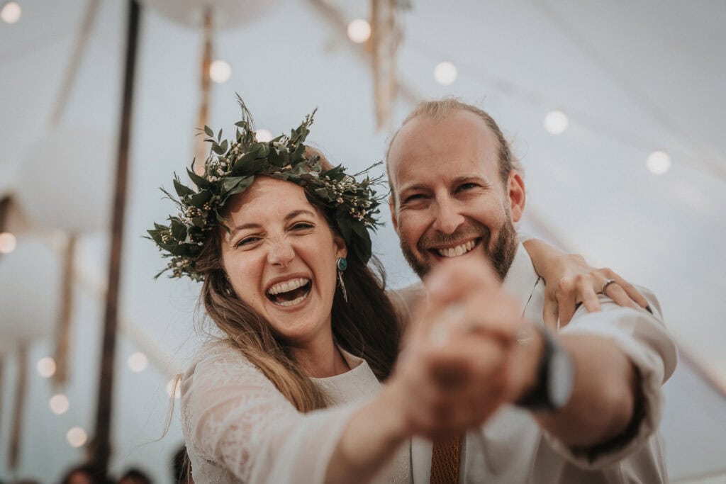 Bride and groom share a joyful first dance under a beautifully lit marquee, with the bride wearing a flower crown and both radiating happiness.