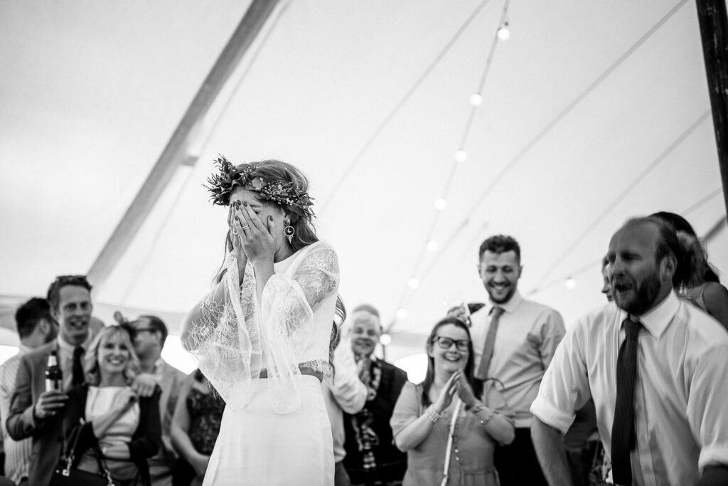 Black and white photo capturing a joyful moment at a wedding reception under a marquee tent. The bride, wearing a floral crown and a lace dress, is overwhelmed with emotion, covering her face with her hands as guests cheer and smile around her. The groom, standing to the right, laughs warmly, surrounded by family and friends in a celebratory atmosphere.