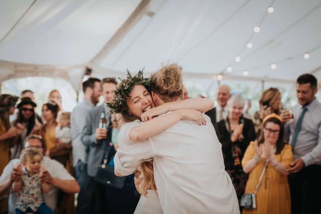 An intimate moment captured between the bride and groom as they embrace under a beautifully lit marquee, surrounded by smiling guests celebrating their special day.