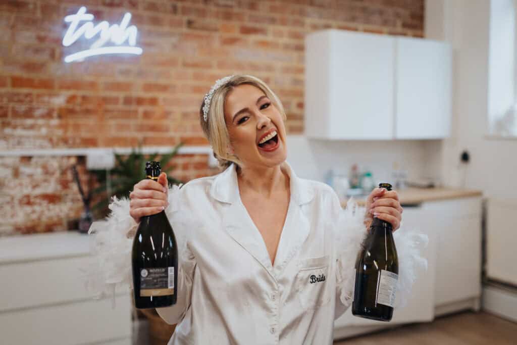 Bride holding two champagne bottles and smiling during fun morning preparations at Thorpe Garden winter wedding, wearing satin pyjamas with “bride” embroidered.