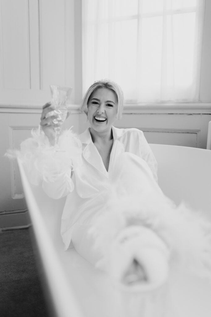 Bride laughing and holding a glass while relaxing in a bathtub during morning preparations at Thorpe Garden winter wedding, wearing satin pyjamas and bridal slippers.