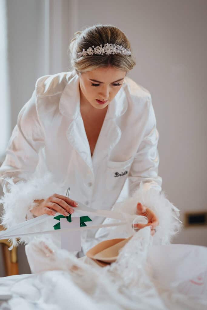 Bride holding hanger and arranging her wedding dress during morning preparations at Thorpe Garden winter wedding, wearing satin pyjamas with feather details.