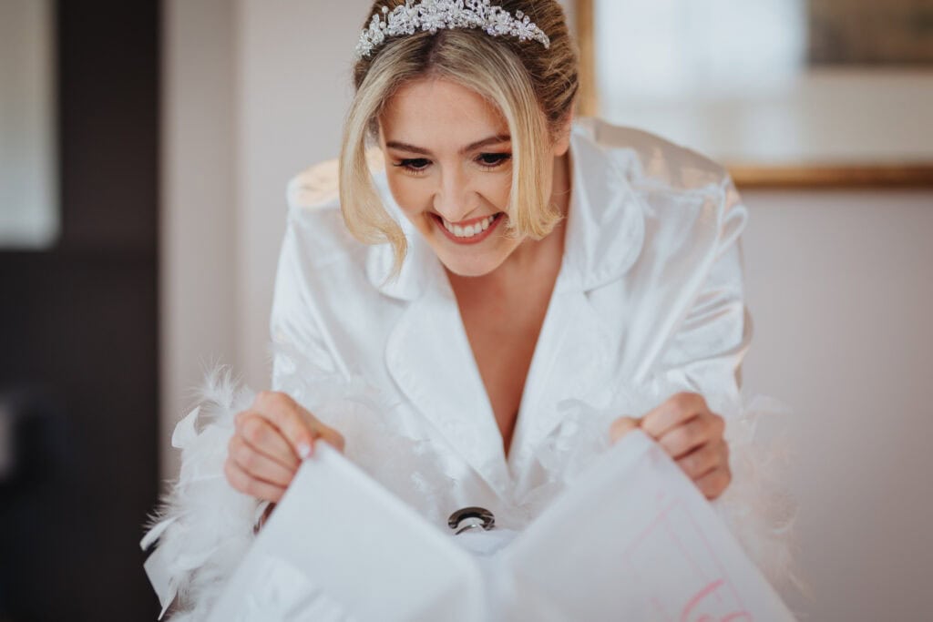 Bride in satin pyjamas with feather details smiles while unpacking her wedding dress during morning preparations at Thorpe Garden winter wedding.