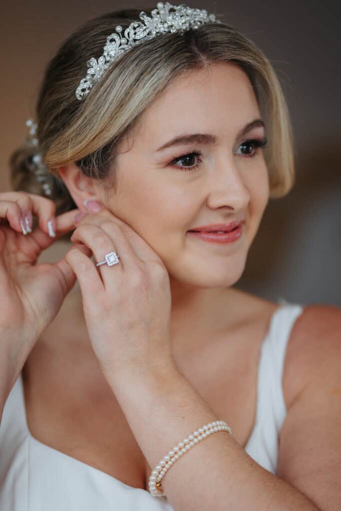 Close-up of bride smiling while putting on earrings during morning preparations at Thorpe Garden winter wedding, wearing a pearl bracelet and sparkling tiara.