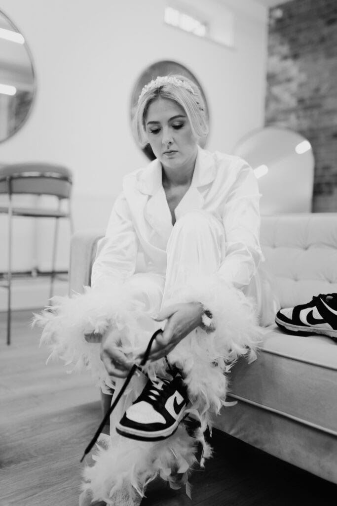 Bride ties black and white trainers while wearing feather-trimmed satin pyjamas during fun morning preparations at Thorpe Garden winter wedding.
