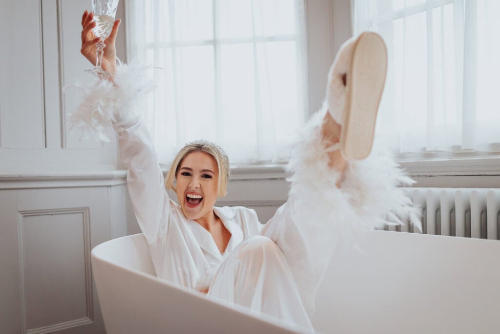Bride laughing and raising a glass while sitting in a bathtub during fun morning preparations at Thorpe Garden winter wedding, wearing satin pyjamas with feather cuffs.